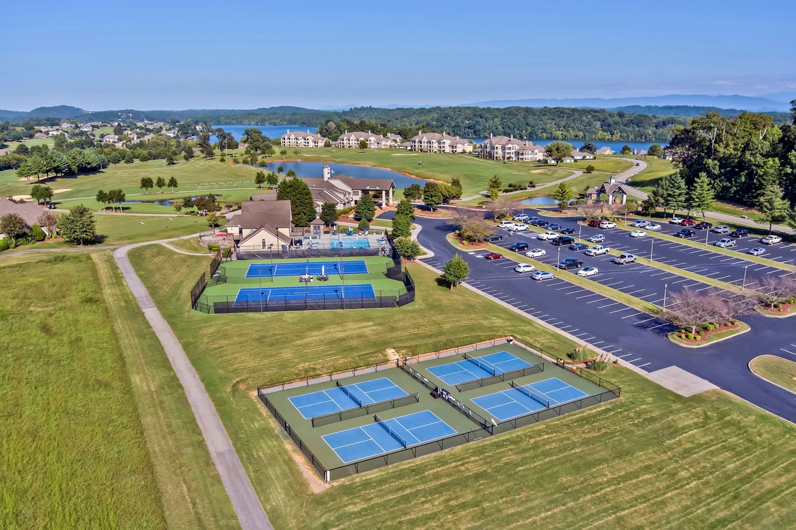 Aerial view of tennis courts, parking lot, and clubhouse beside a lake and green fields.