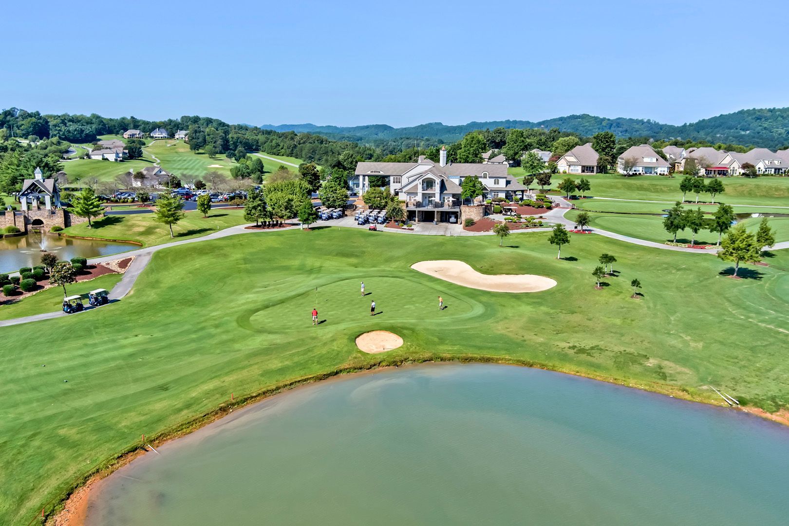 Golf course with green grass, lake, clubhouse, and homes in the background under a blue sky.