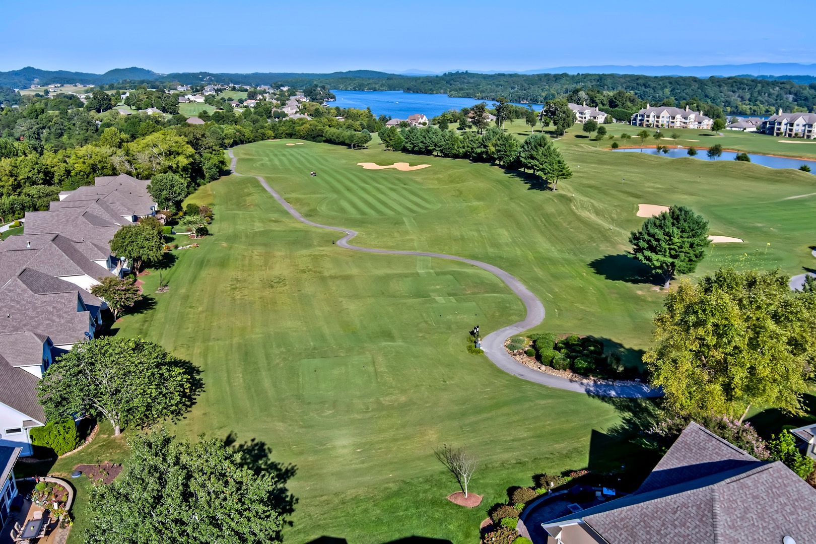 Aerial view of a green golf course with a winding path, houses, and a lake in the distance under a blue sky.