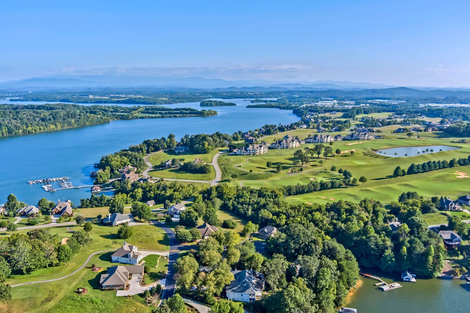 Aerial view of a lake and green landscape with houses, trees, and a golf course under a blue sky.