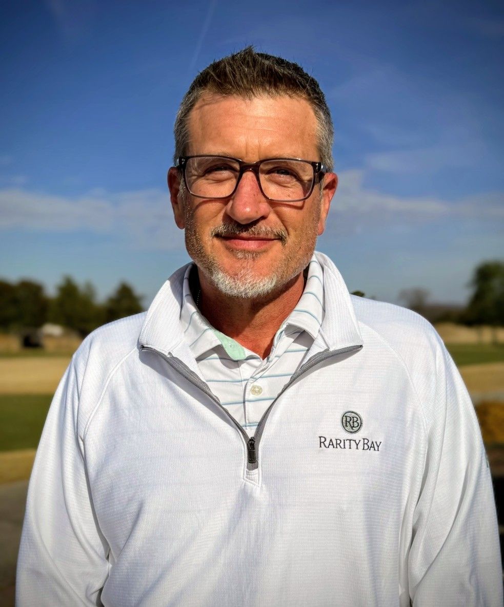 Tennis player in white shorts and visor swings racket on green court, shadow visible.