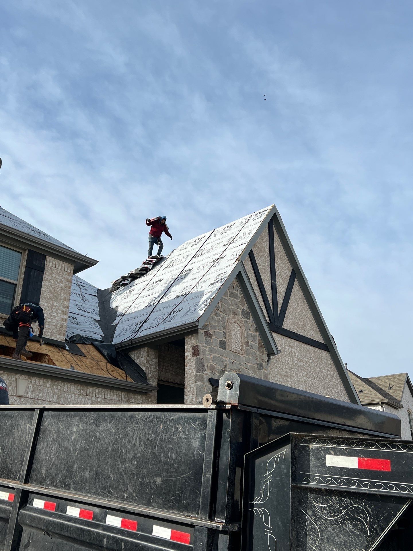 Roofers working on a residential roof, removing old shingles and replacing them. Dumpster in foreground.