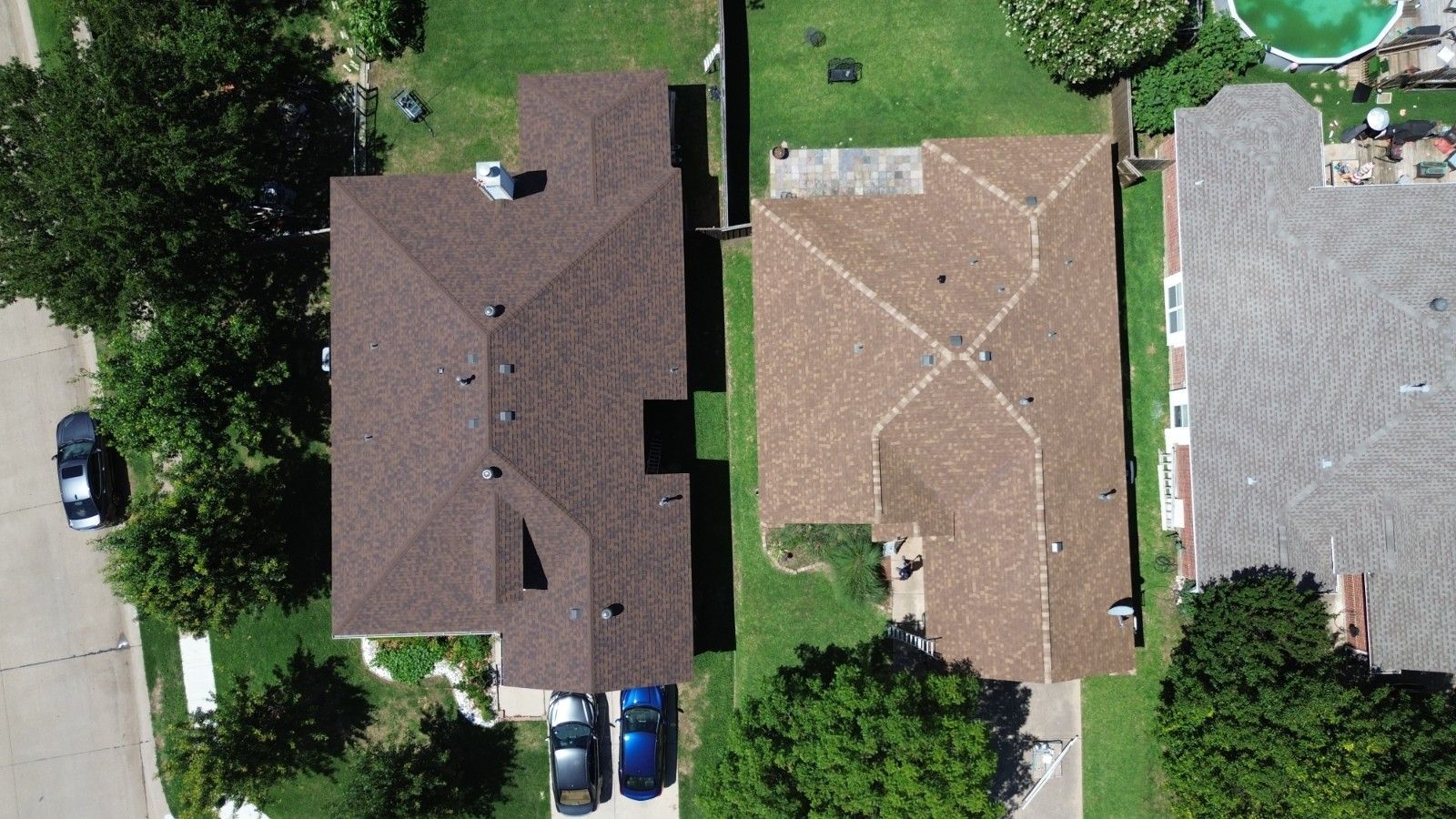 Two houses with brown roofs side-by-side on green lawns, seen from above. Cars parked in front of one.