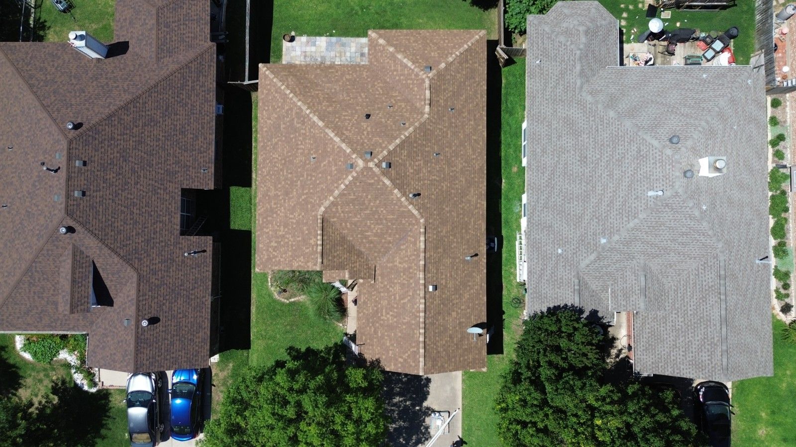 Overhead view of three houses with brown, grey and tan roofs, surrounded by green grass and trees.