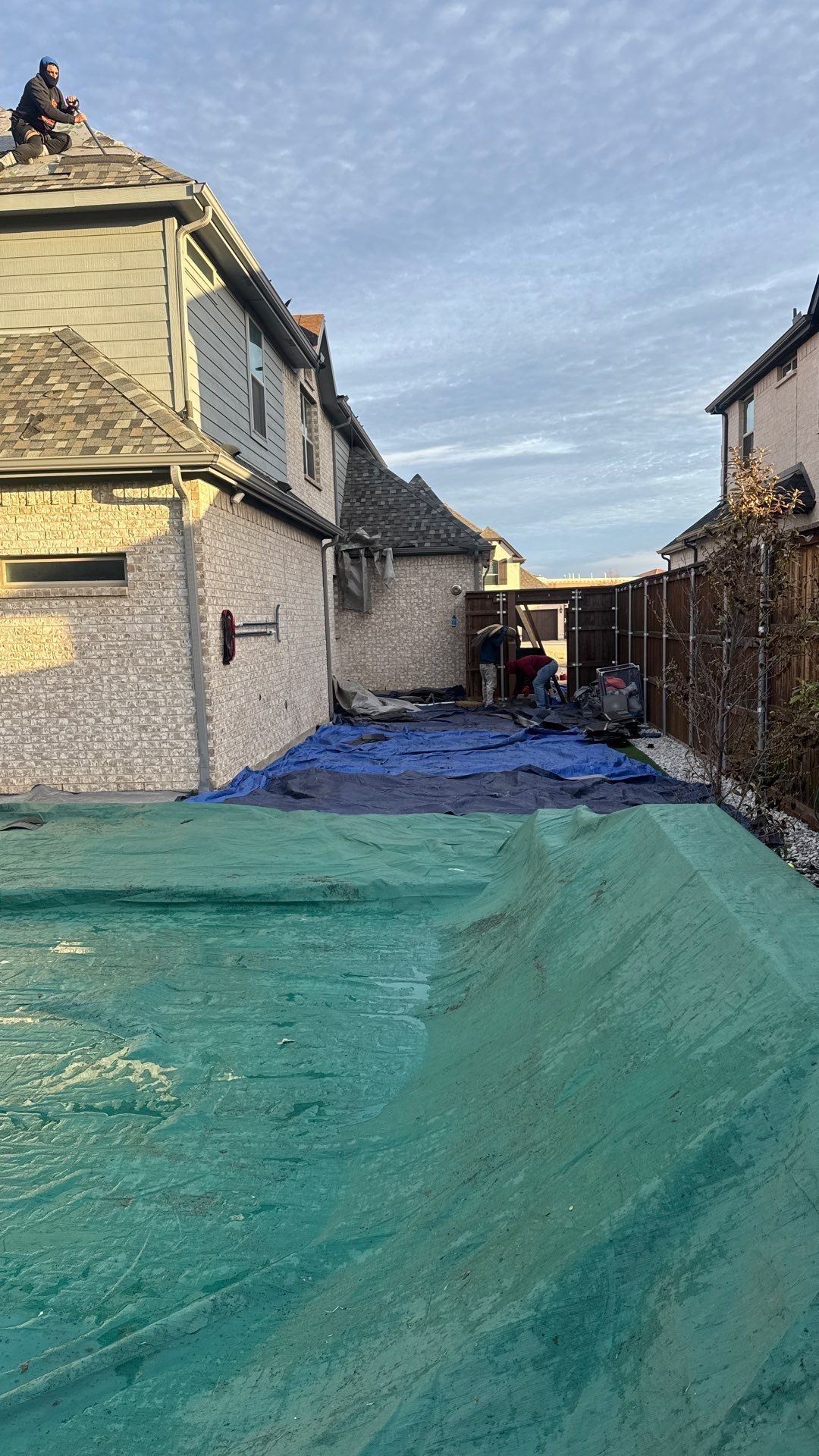 Workers on a rooftop and in a yard with blue tarps. Brick building and green ground covering.
