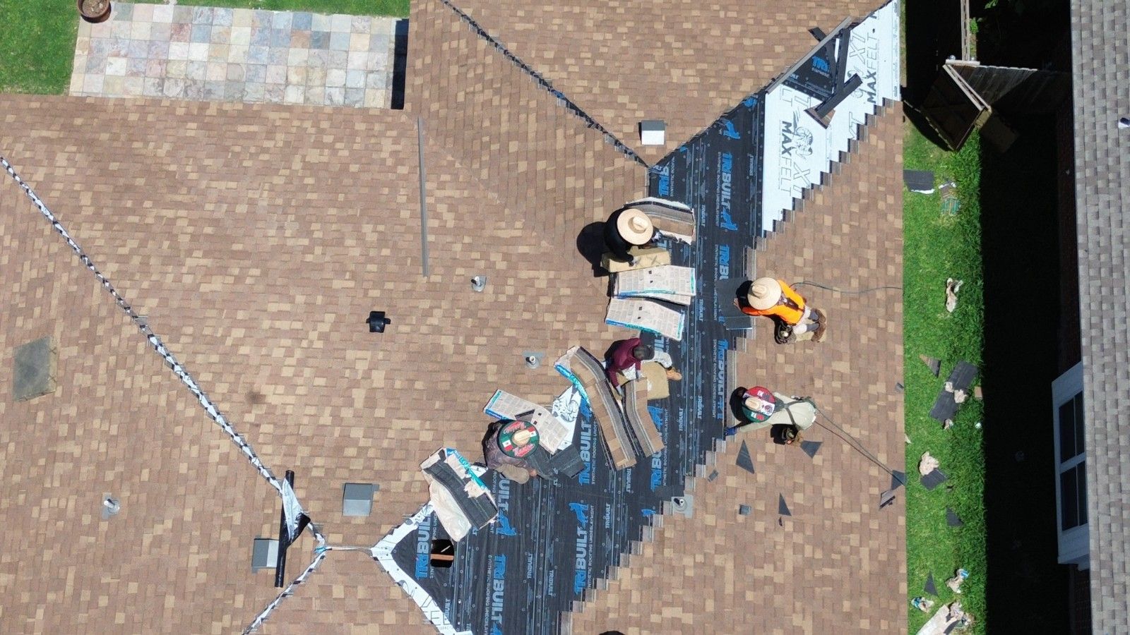 Aerial view of roofers installing new shingles on a brown shingled roof, next to a green lawn.