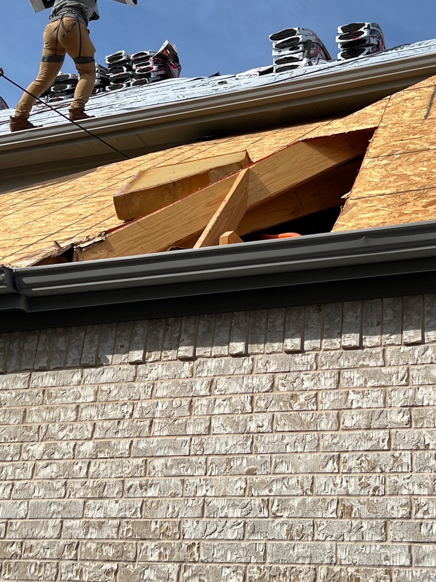 Roofer on a roof removes damaged roofing; new plywood structure revealed. Brick building, blue sky background.