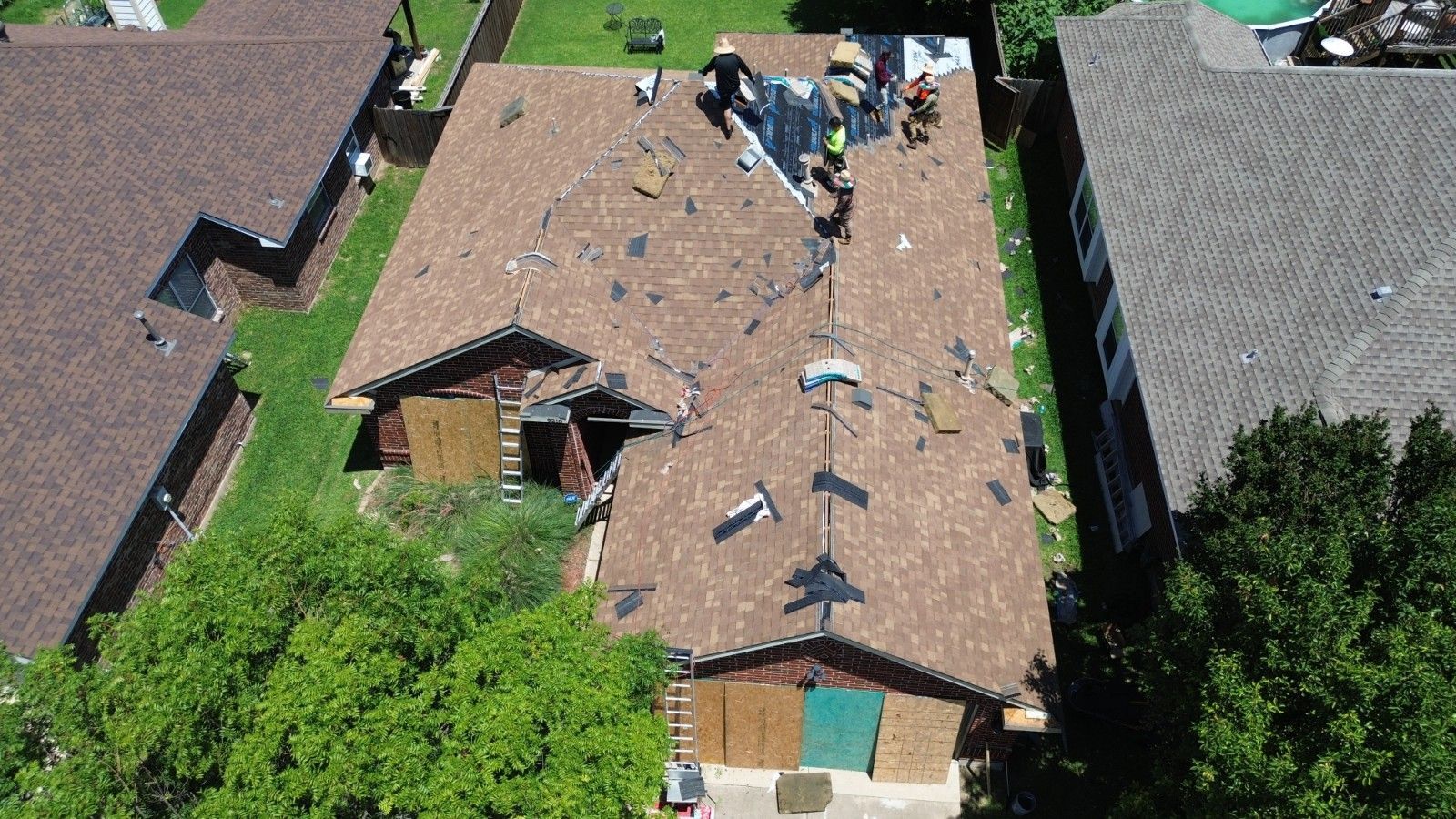 Roof of a house damaged with people on it, working. Brown shingles, boarded-up sections, green grass.