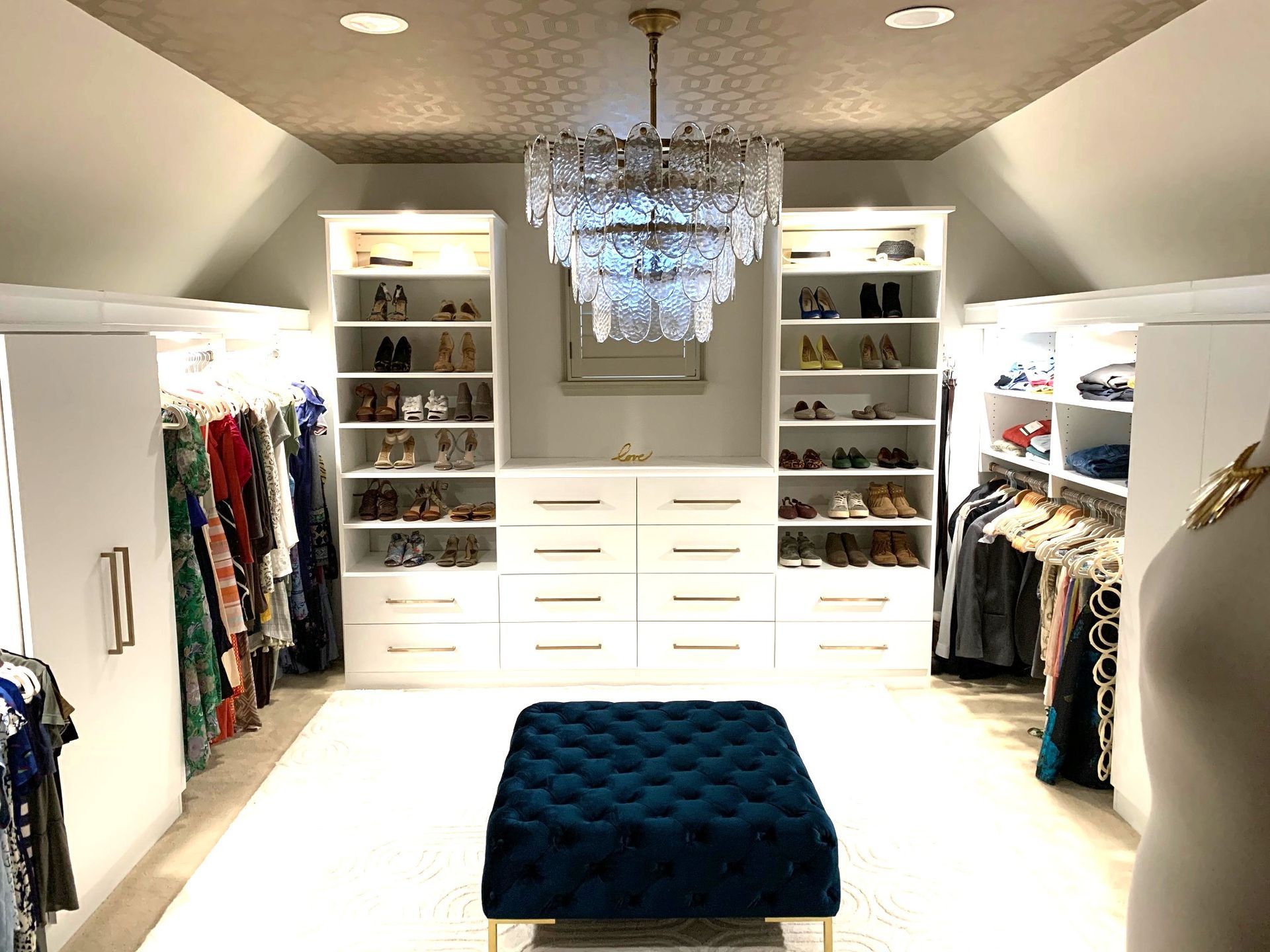 A large walk in closet with a blue ottoman and a chandelier.