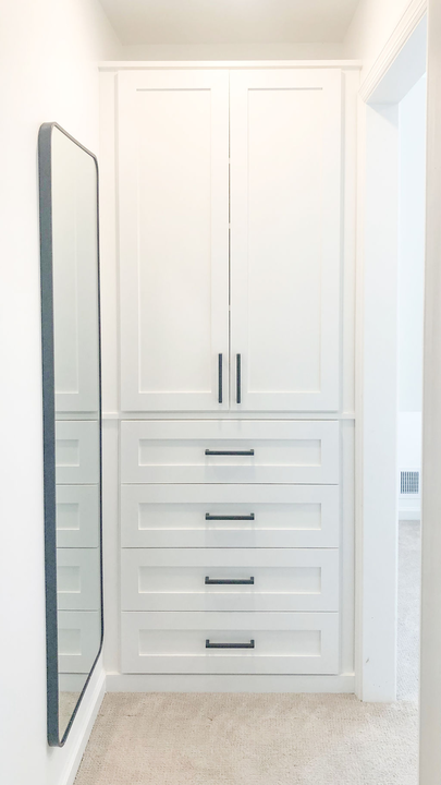 A walk in closet with white cabinets and drawers and a large mirror.