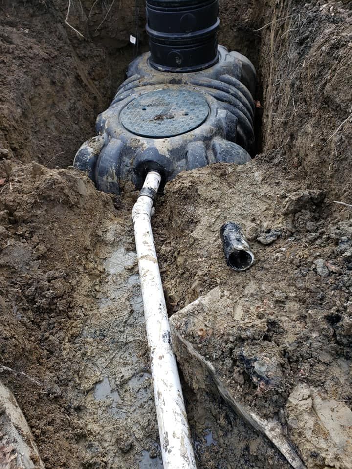 Black septic tank in trench, white pipe leading to it. Brown dirt surrounds.