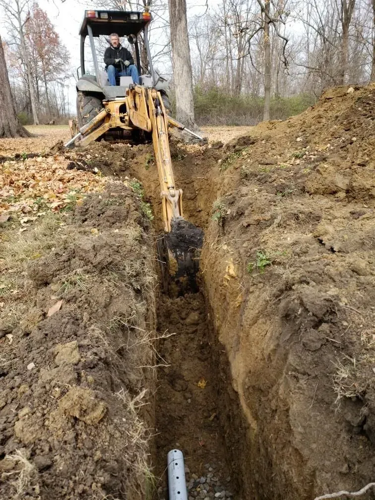 Tractor digging a trench for a pipe in a yard; a person is operating the machinery.