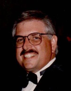 A man with glasses and a mustache is wearing a tuxedo and smiling.