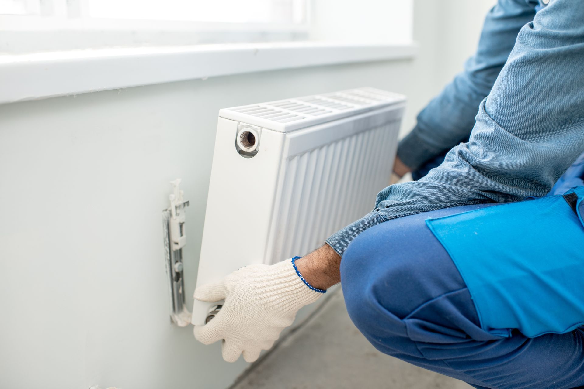 A worker in work gloves installs a white radiator on a wall under a window.
