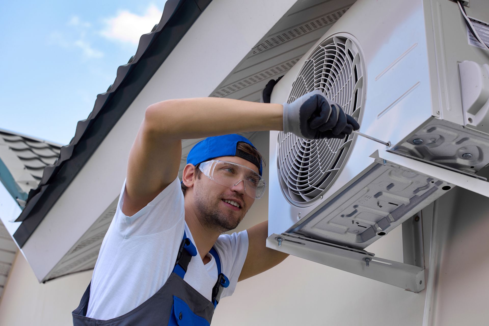 A technician in a white shirt and blue work overalls wears safety goggles while repairing an outdoor air conditioner.