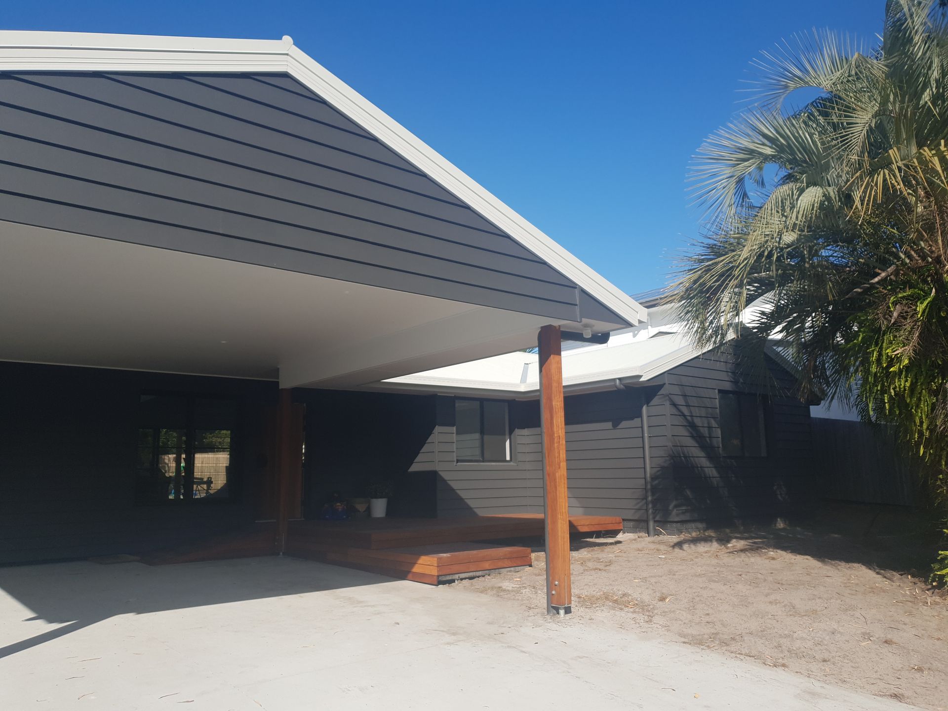 Modern house carport with dark siding, white roof eaves, wooden post, and palm tree in front