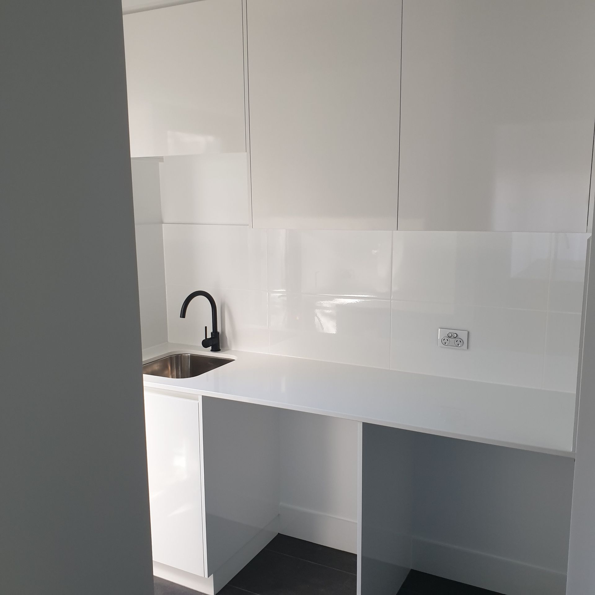 Modern white kitchenette with sink, black faucet, glossy backsplash, and open cabinet space