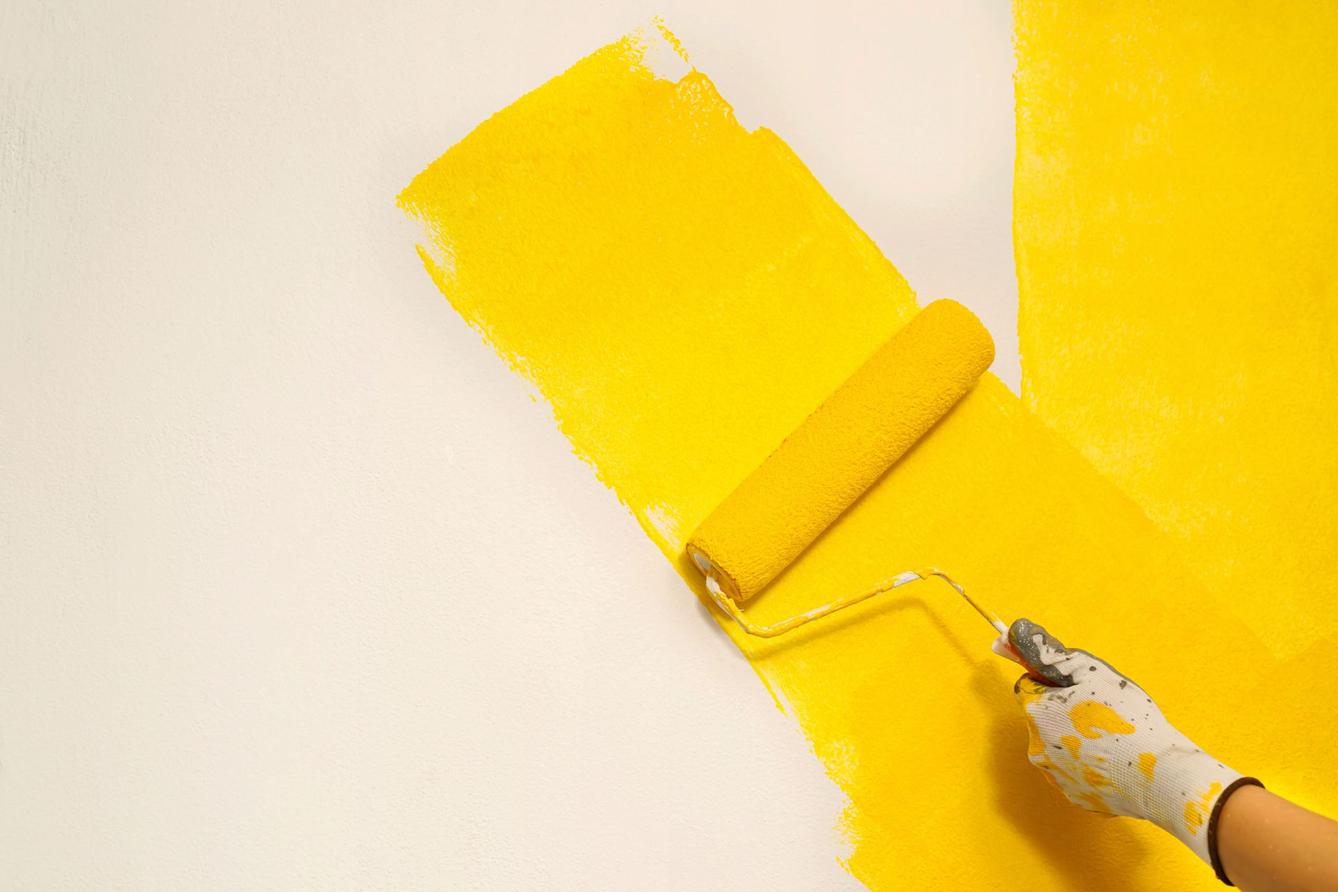 Persona con guanto da lavoro che dipinge una parete bianca con un rullo di vernice gialla.