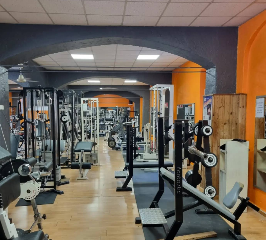 Interno di una palestra con attrezzi per il sollevamento pesi, una panca e pesi. Pareti arancioni e nere, pavimento in legno.