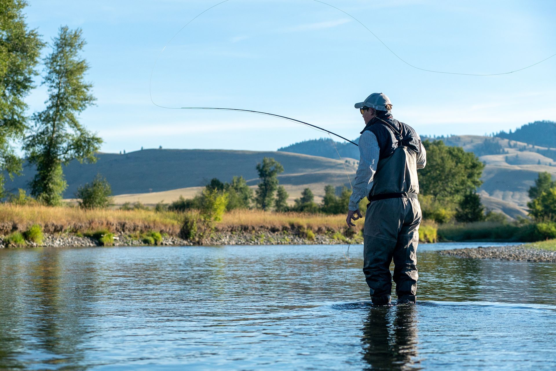 A man is fishing in a river with mountains in the background.