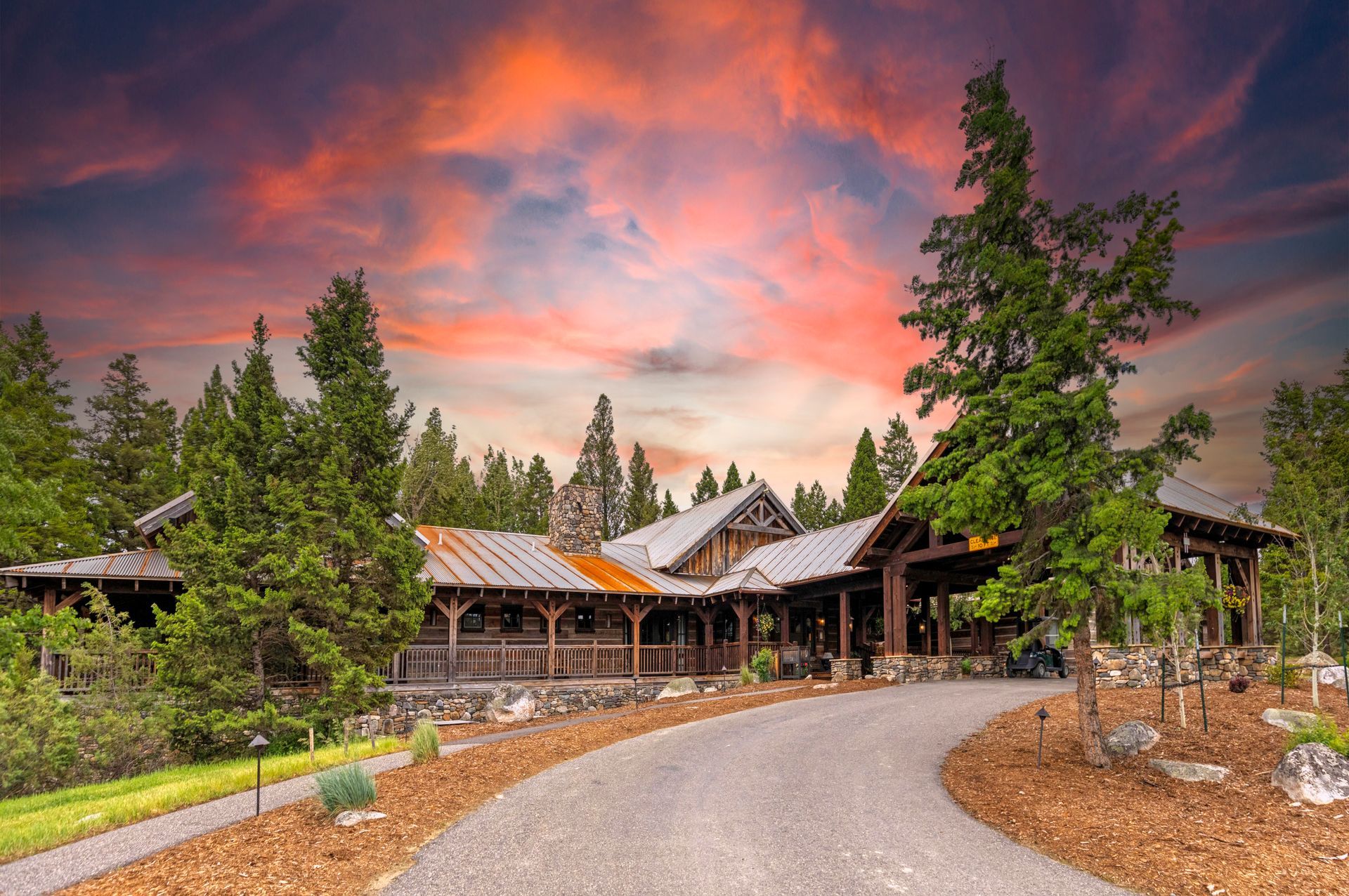 A large house with a driveway leading to it is surrounded by trees and a sunset in the background.
