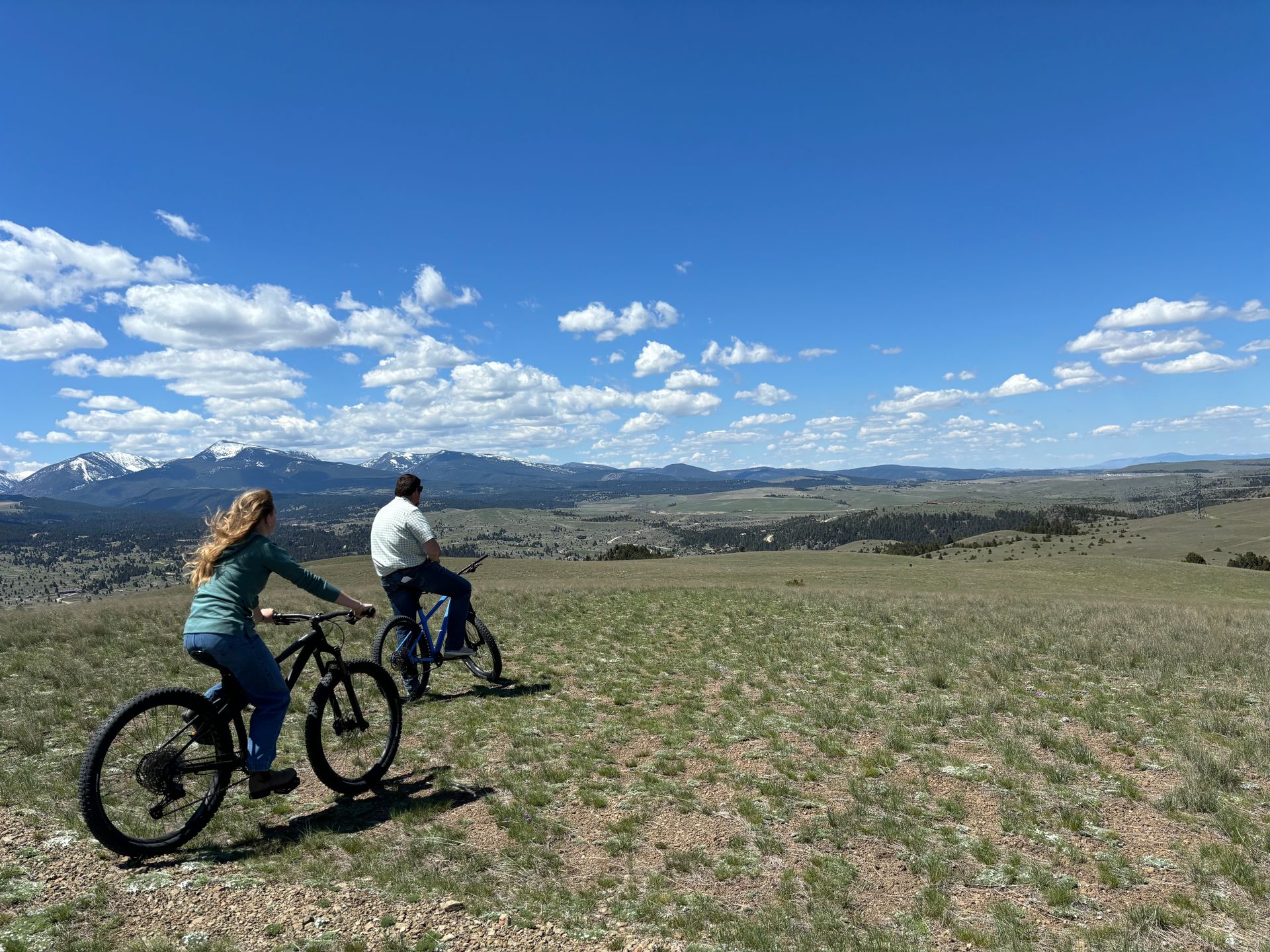 A man and a woman are riding bicycles on top of a grassy hill.