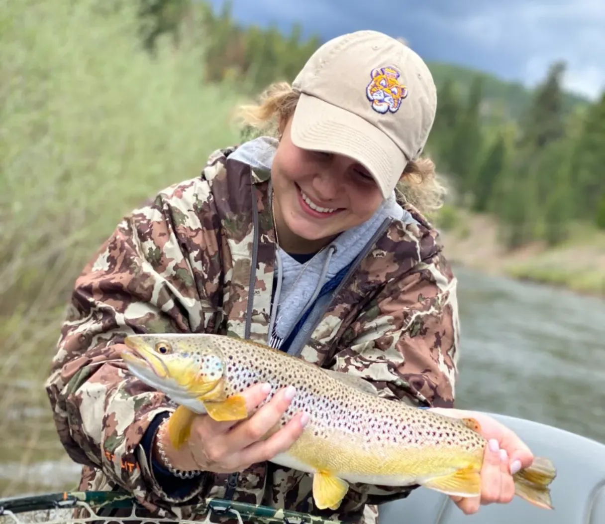 A woman in a hat is holding a brown trout in her hands.