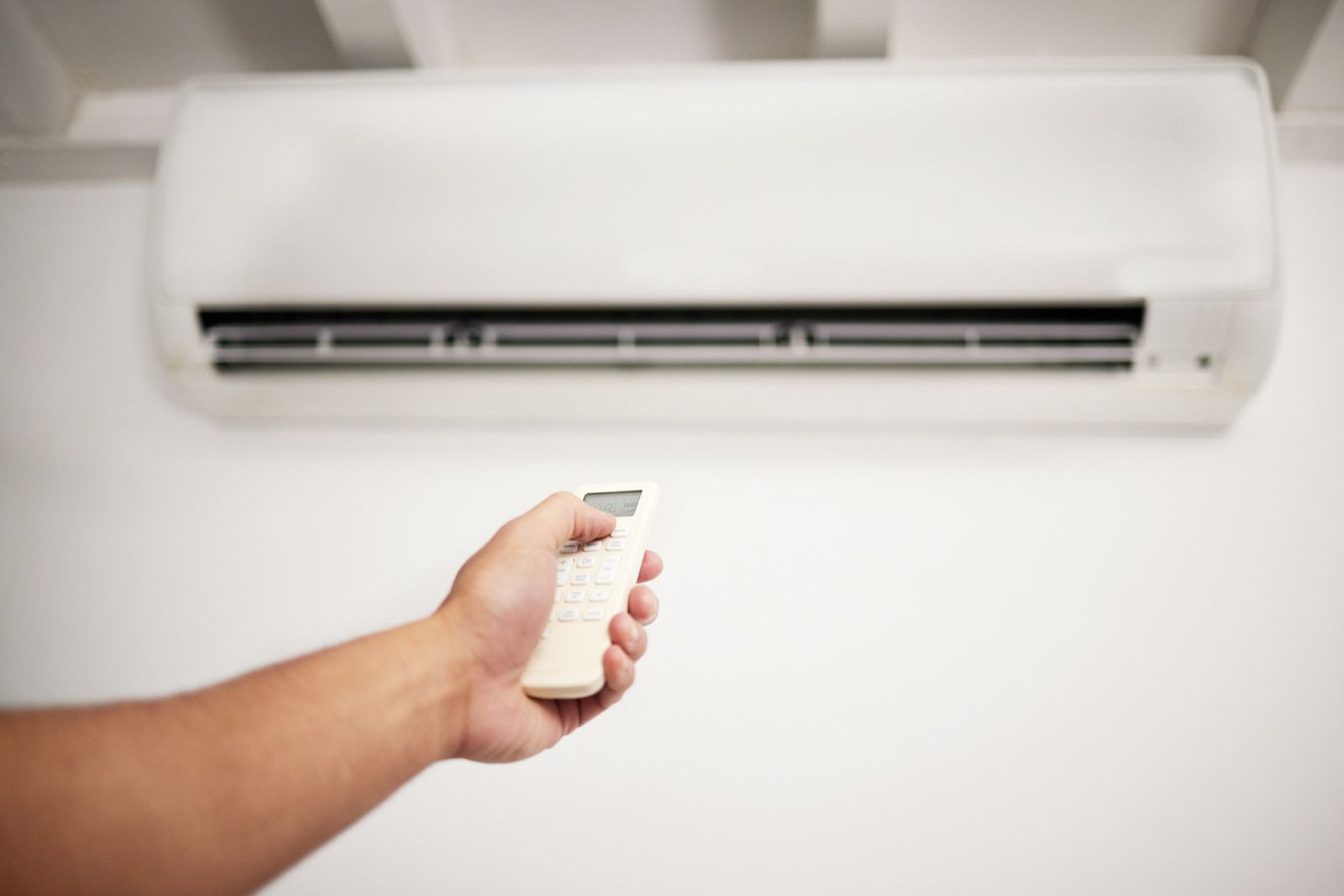 A hand holding a remote control aimed at a white wall-mounted air conditioner.