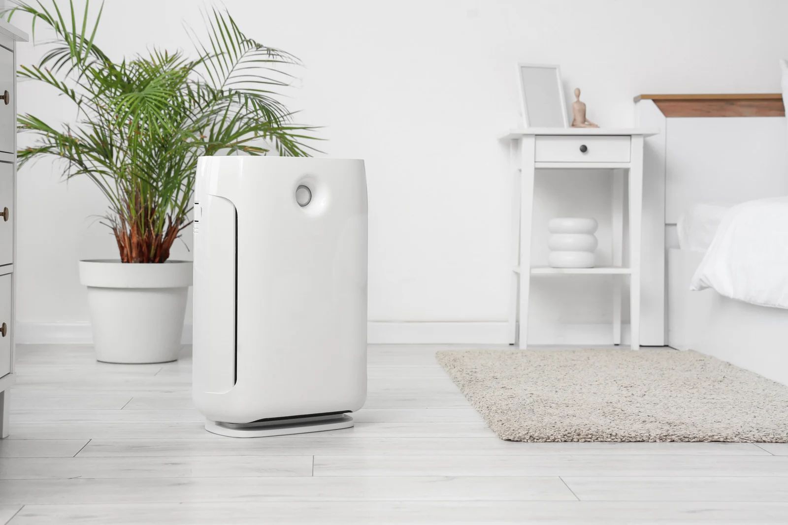 White air purifier in a bedroom with a potted plant, nightstand, and rug.