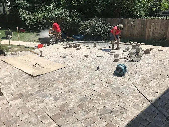 Two men are working on a brick patio in a backyard.