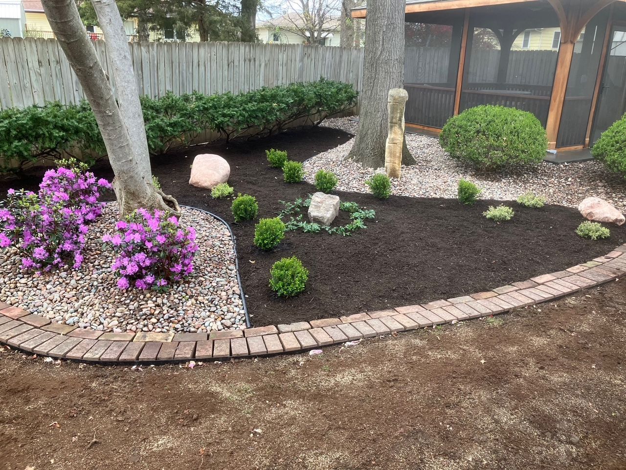 A landscaped garden bed with purple flowers, dark mulch, and a brick border.