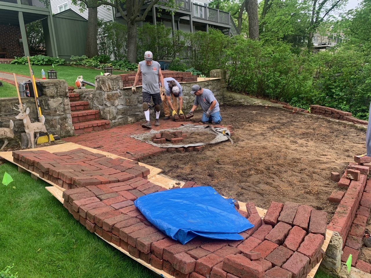 Three workers laying brick patio in a yard, surrounded by greenery and an old stone structure.