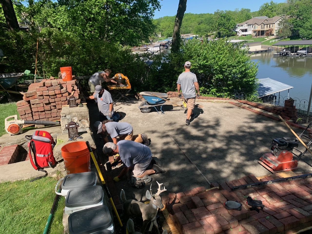 Workers laying brick patio near a lake. Several men, tools, and bricks. Sunny day.