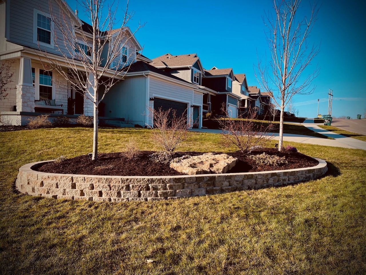 A landscaped yard with a stone retaining wall, trees, and houses under a blue sky.
