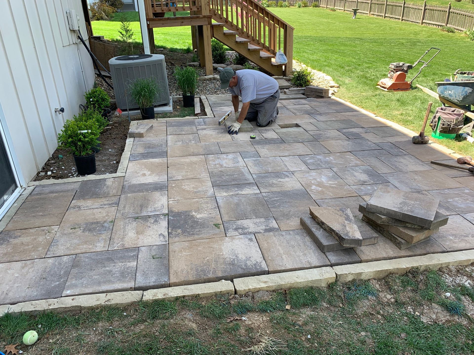 Man kneeling, laying patio pavers near a house, grass, and deck.