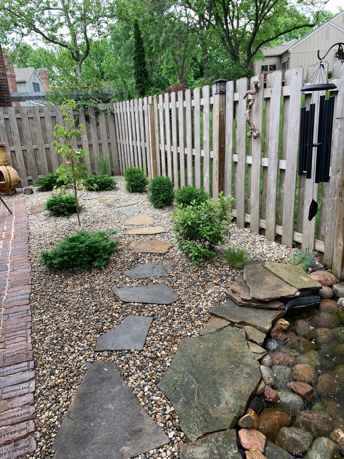 A small backyard garden with stepping stones, shrubs, gravel, and a fence.