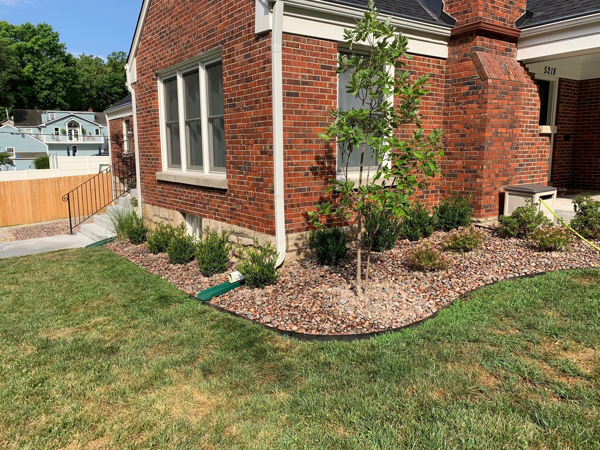 Red brick house with landscaping, including a small tree and shrubs, edged with brown stones.
