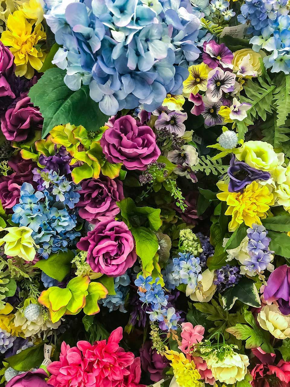 Colorful Mixed Floral Arrangement Featuring Blue Hydrangeas, Purple Roses, Yellow Blossoms, And Lush Green Foliage Filling The Frame
