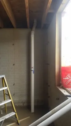A white vertical plumbing pipe runs from the concrete floor to the wooden ceiling joists in an unfinished basement.