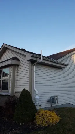 A white exterior pipe for a radon mitigation system runs up the side of a beige-sided house near a gas meter.