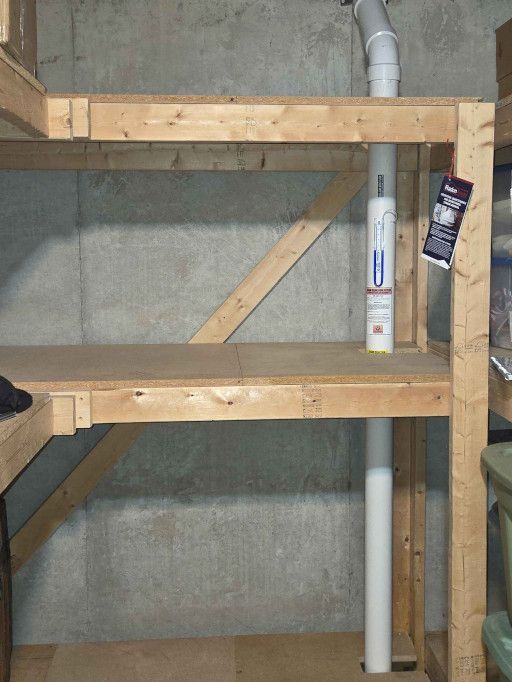A radon mitigation pipe runs vertically through a wooden storage shelf against a concrete basement wall.