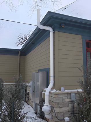 A white radon mitigation pipe runs along the side of a tan and stone-sided building in the snow.