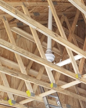 A white PVC plumbing vent pipe runs vertically through wooden roof trusses in an unfinished attic space.
