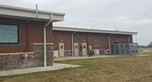 A single-story brick building with exterior doors and an HVAC unit on a paved walkway next to a field.