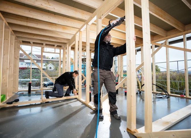 Two workers framing a wooden house interior under construction, one standing and one kneeling.