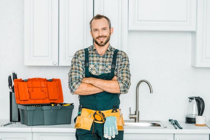 Handyman in a kitchen, arms crossed, wearing a tool belt beside an open toolbox and sink.