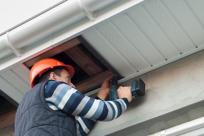 Worker in orange hard hat using a drill under the roof eaves