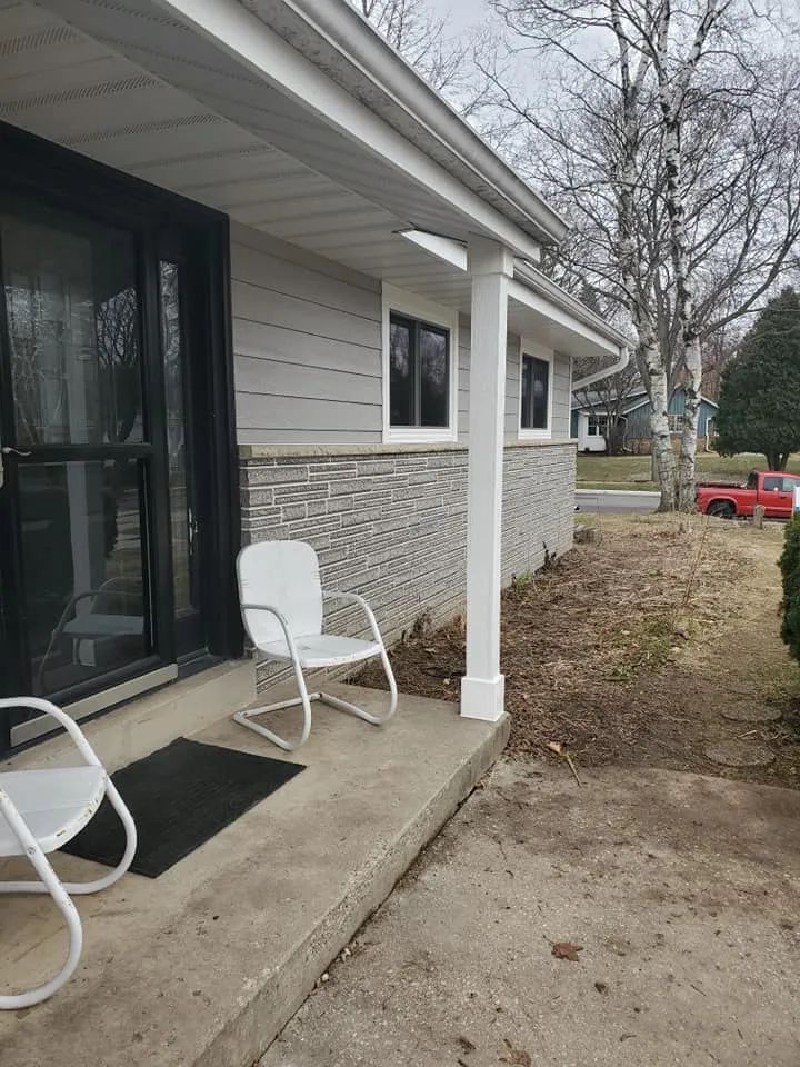 A house with a porch. Two white chairs sit on a concrete porch, next to a black door, with light gray siding.