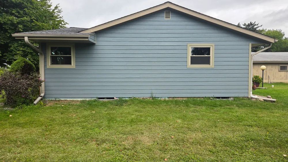 Blue-sided house with green trim and two windows set on a grassy lawn under an overcast sky.