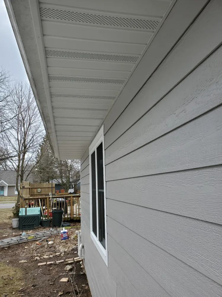 Gray siding and white trim on a house, window on the right. Overhang with vents above.